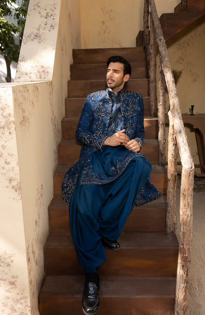 A man wearing a traditional Indian outfit with a blue patterned kurta and matching trousers sits on wooden stairs in an outdoor setting.