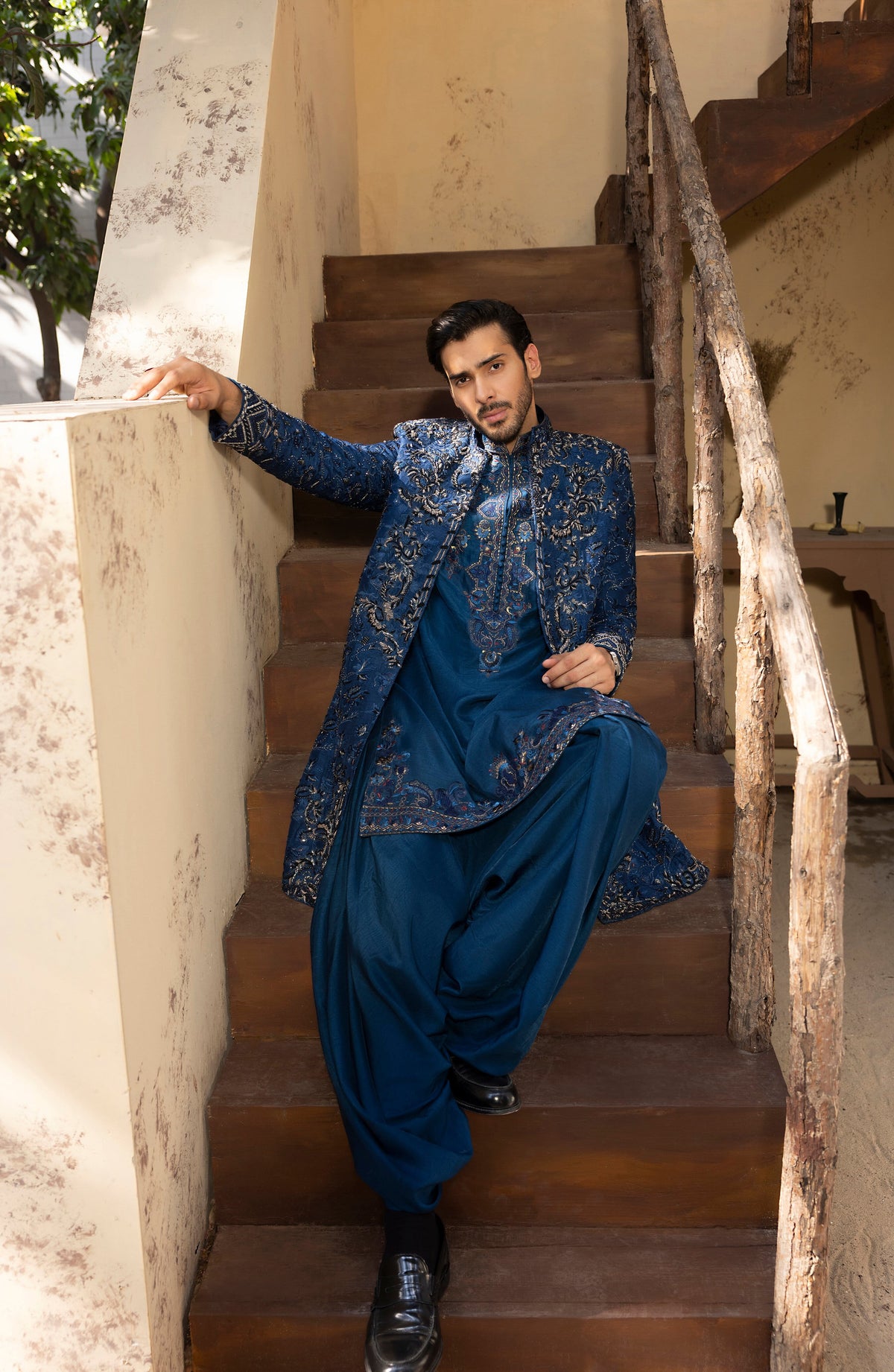 A man wearing a traditional blue embroidered kurta and wide-leg trousers sits on wooden stairs next to a textured wall.
