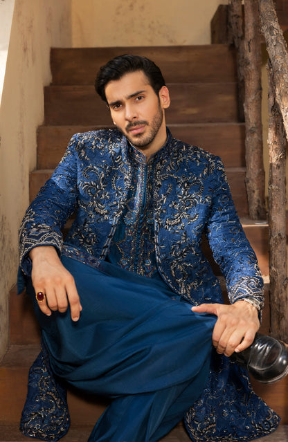 A man wearing a blue and gold embroidered sherwani jacket with matching blue trousers, sitting on rustic wooden stairs.