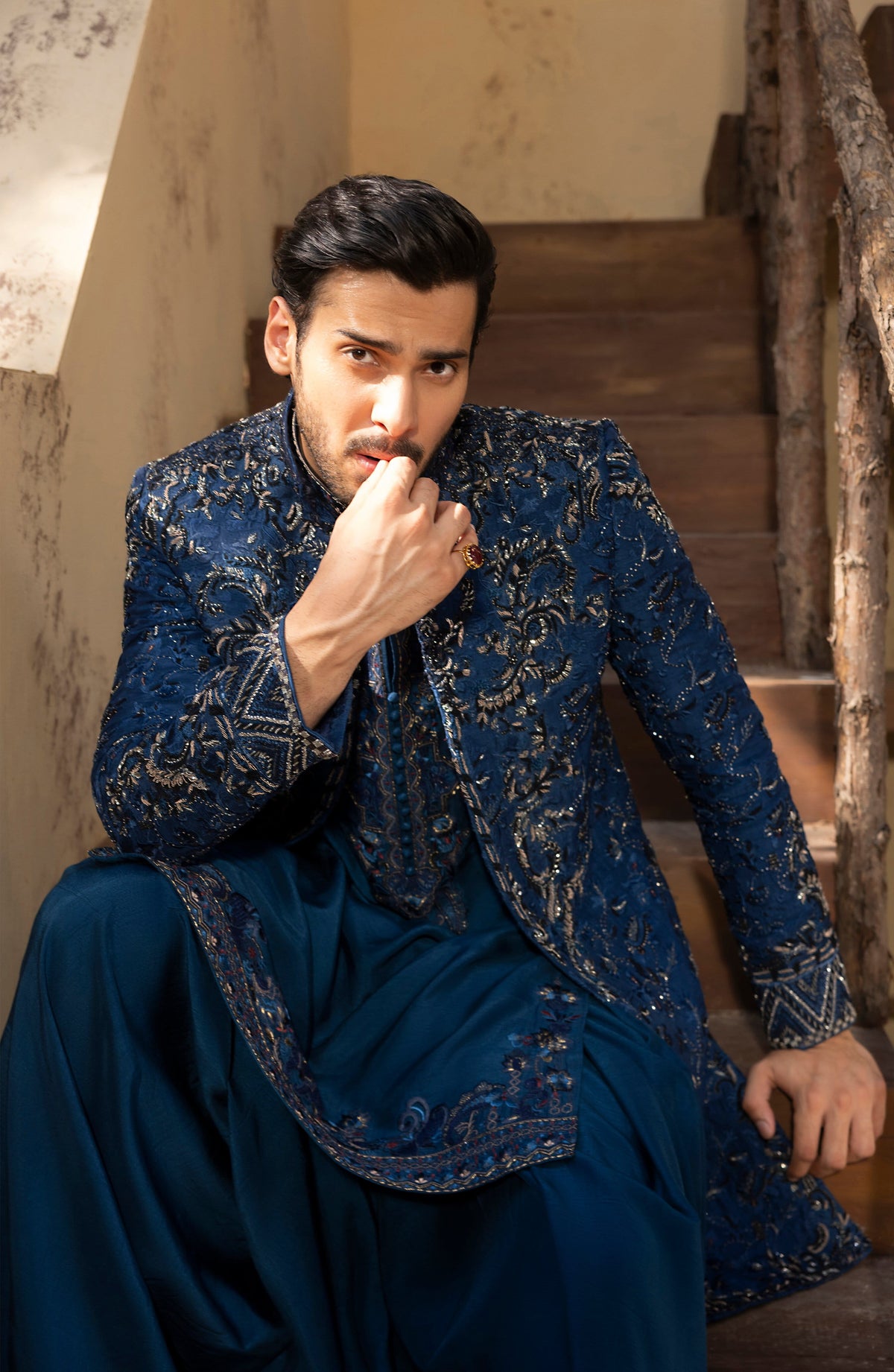 A man wearing an ornate navy blue embroidered sherwani jacket with matching trousers, seated on rustic wooden stairs.