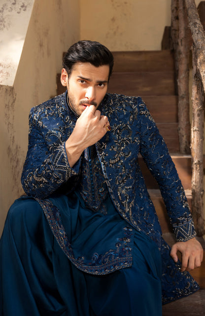 A man wearing an ornate navy blue embroidered sherwani jacket with matching trousers, seated on rustic wooden stairs.