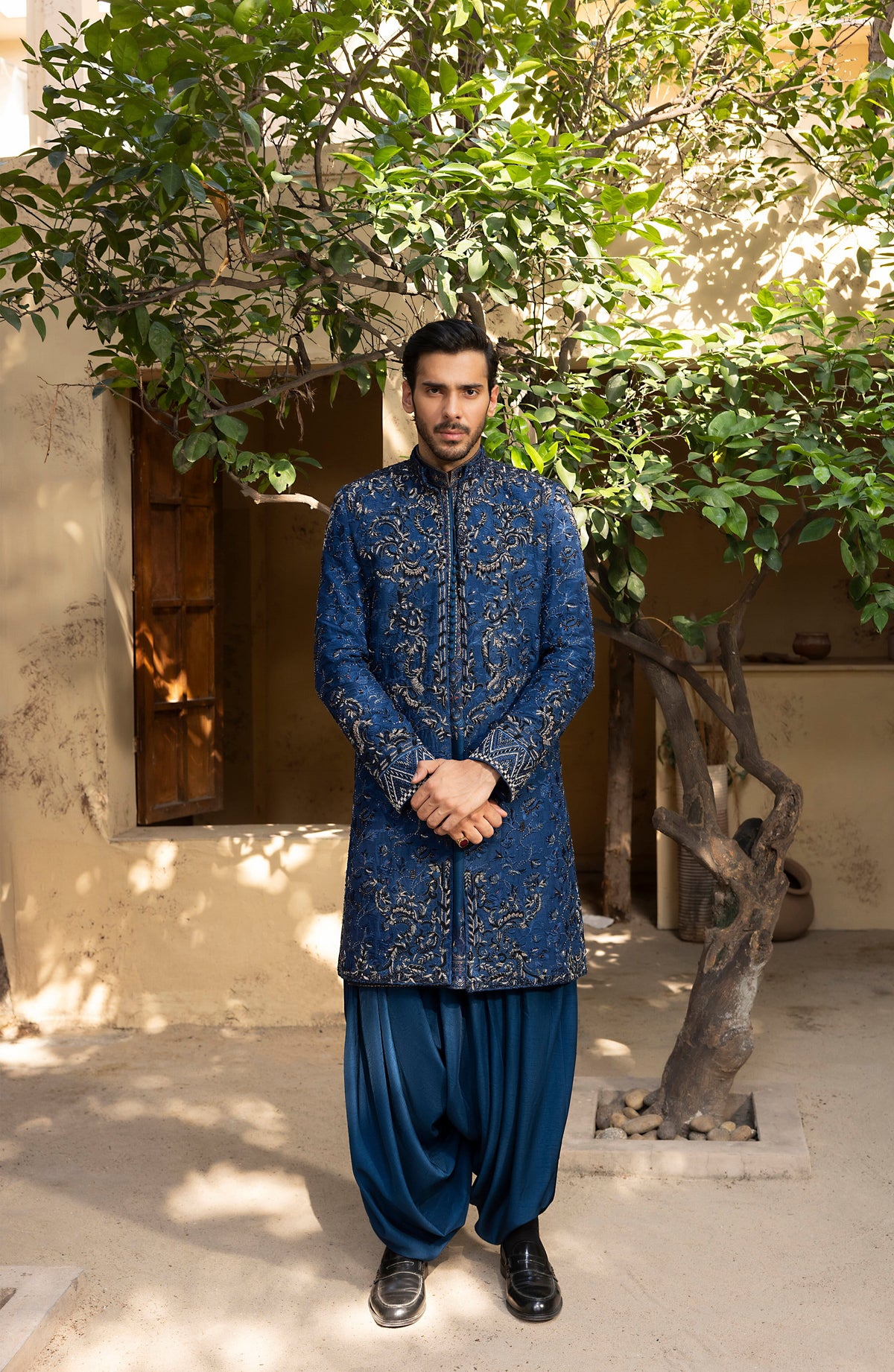 A man wearing a traditional blue embroidered sherwani jacket with matching blue trousers, standing outdoors in front of a beige building and leafy tree.