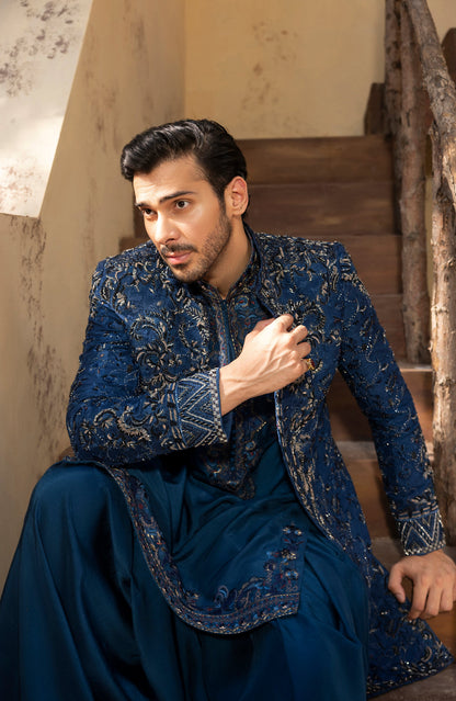 A man wearing a dark blue traditional embroidered kurta and matching trousers, sitting on wooden stairs in a warm-toned interior setting.