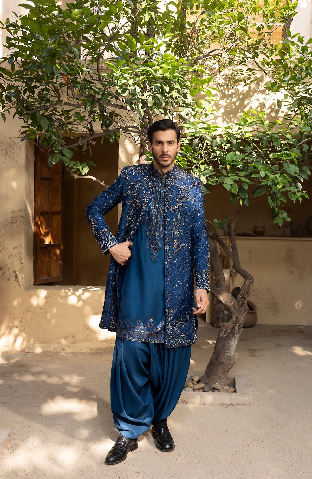 A man wearing a traditional blue embroidered kurta and matching pants stands outdoors in front of a rustic wall and leafy tree.