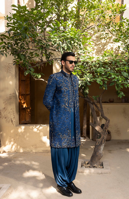 A man wearing a traditional blue embroidered kurta and matching trousers stands outdoors in front of a tree and beige building wall.
