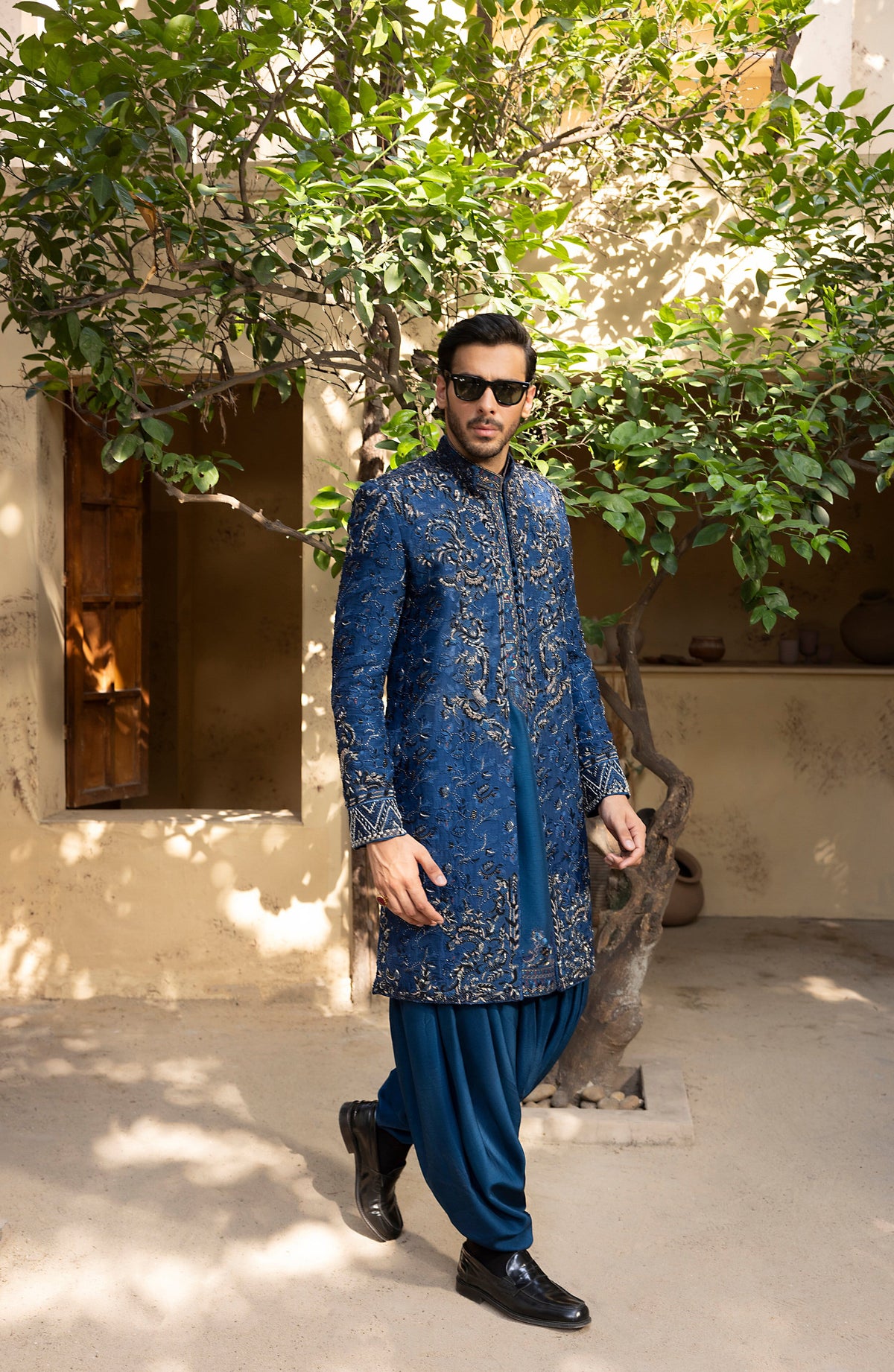 A man wearing a blue embroidered sherwani with gold motifs and matching blue pants, walking outdoors in front of a beige building and green trees.