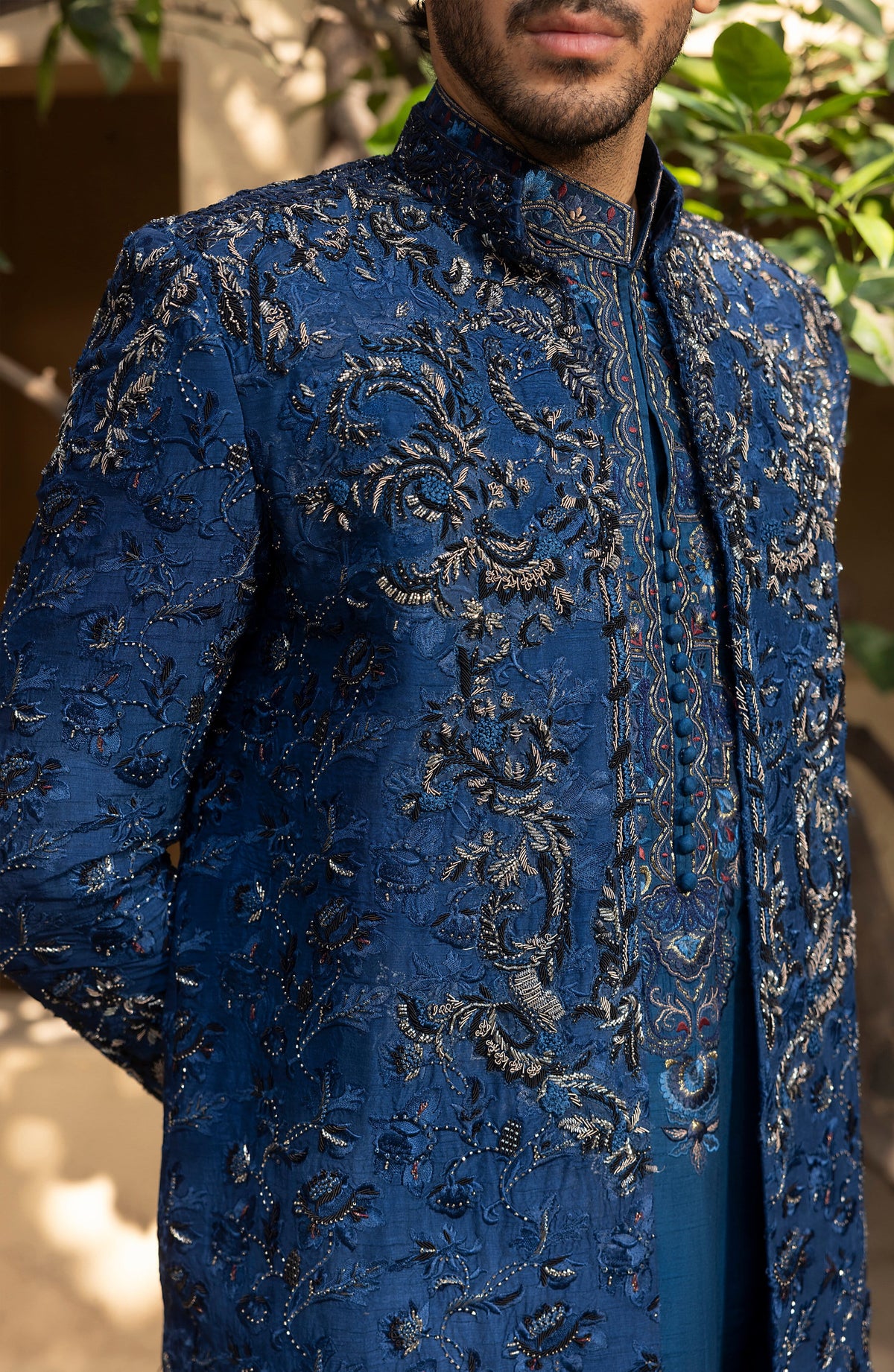 A close-up of a man wearing a rich blue traditional embroidered jacket with intricate metallic silver floral embroidery, set against a soft-focus background of foliage and warm architectural elements.