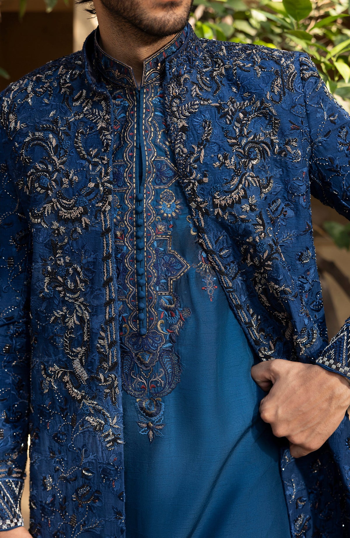 A close-up of a man wearing a royal blue embroidered sherwani jacket over a matching blue kurta, with ornate metallic embroidery and a mandarin collar.