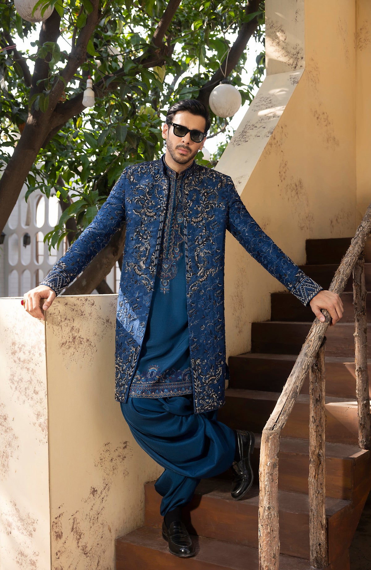 A man wearing a traditional blue embroidered sherwani jacket over a matching blue kurta and trousers, posing on an outdoor wooden staircase.