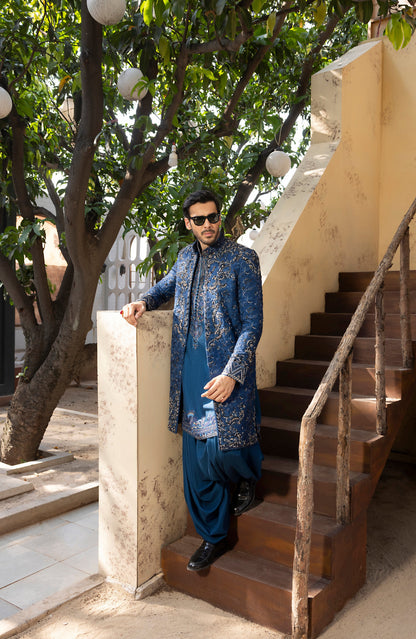 A man wearing a blue patterned sherwani and matching trousers stands on outdoor wooden stairs next to a beige wall and leafy trees.
