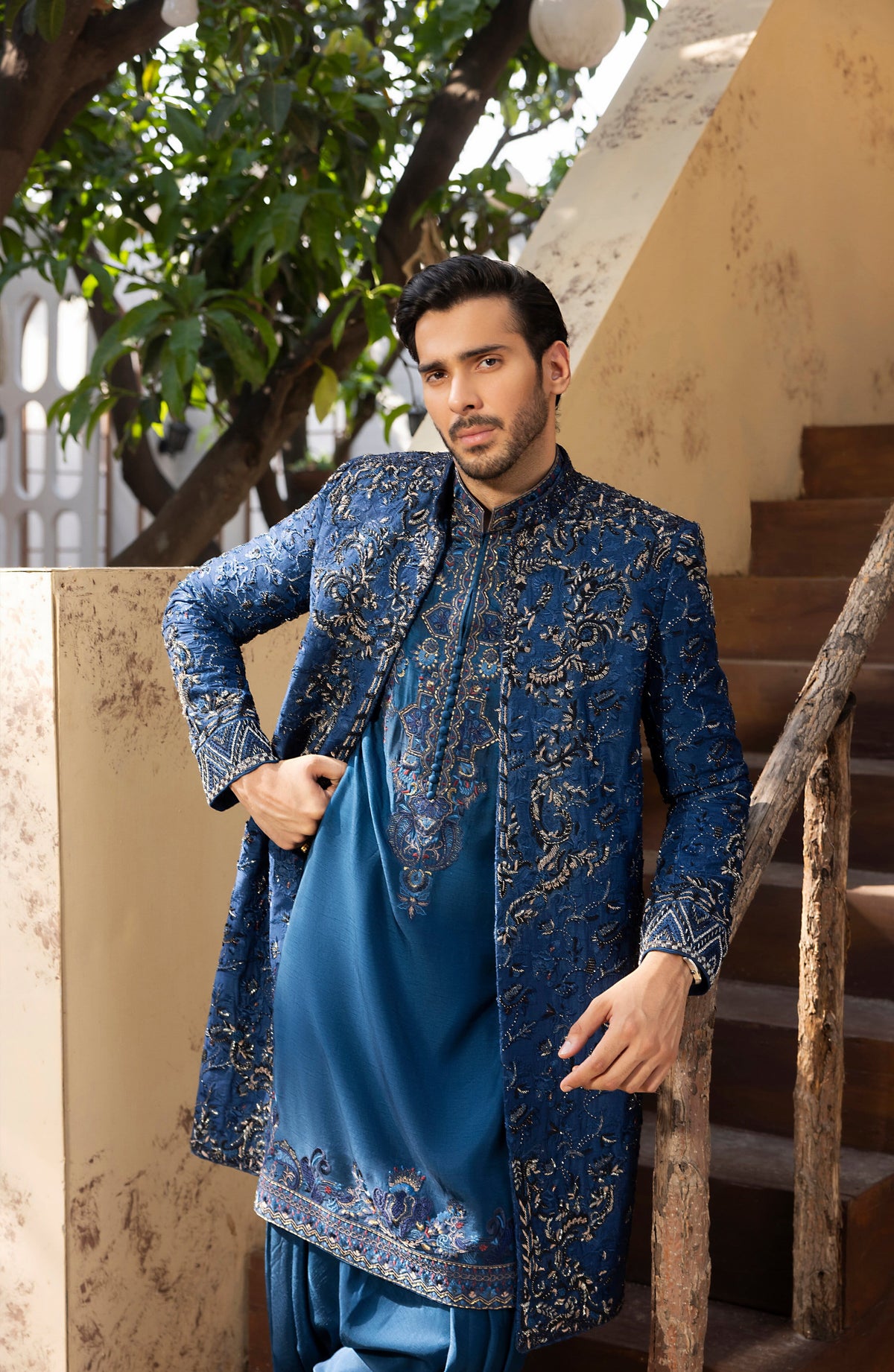 A man wearing a traditional Indian outfit consisting of a blue kurta with gold embroidery and a matching blue jacket, standing in front of a staircase.