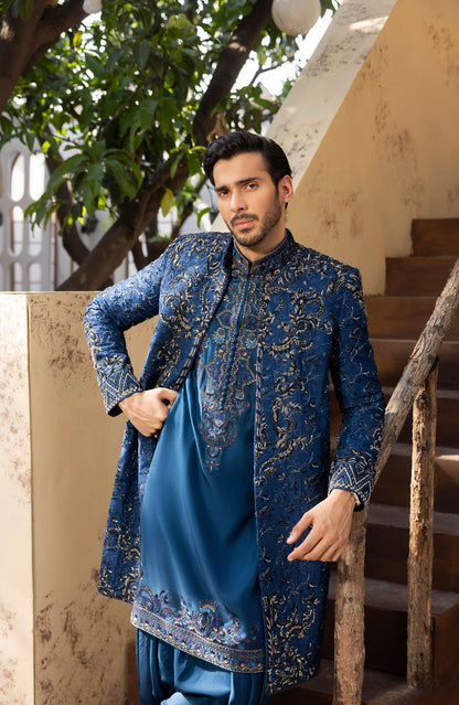A man wearing a traditional Indian outfit consisting of a blue kurta with gold embroidery and a matching blue jacket, standing in front of a staircase.