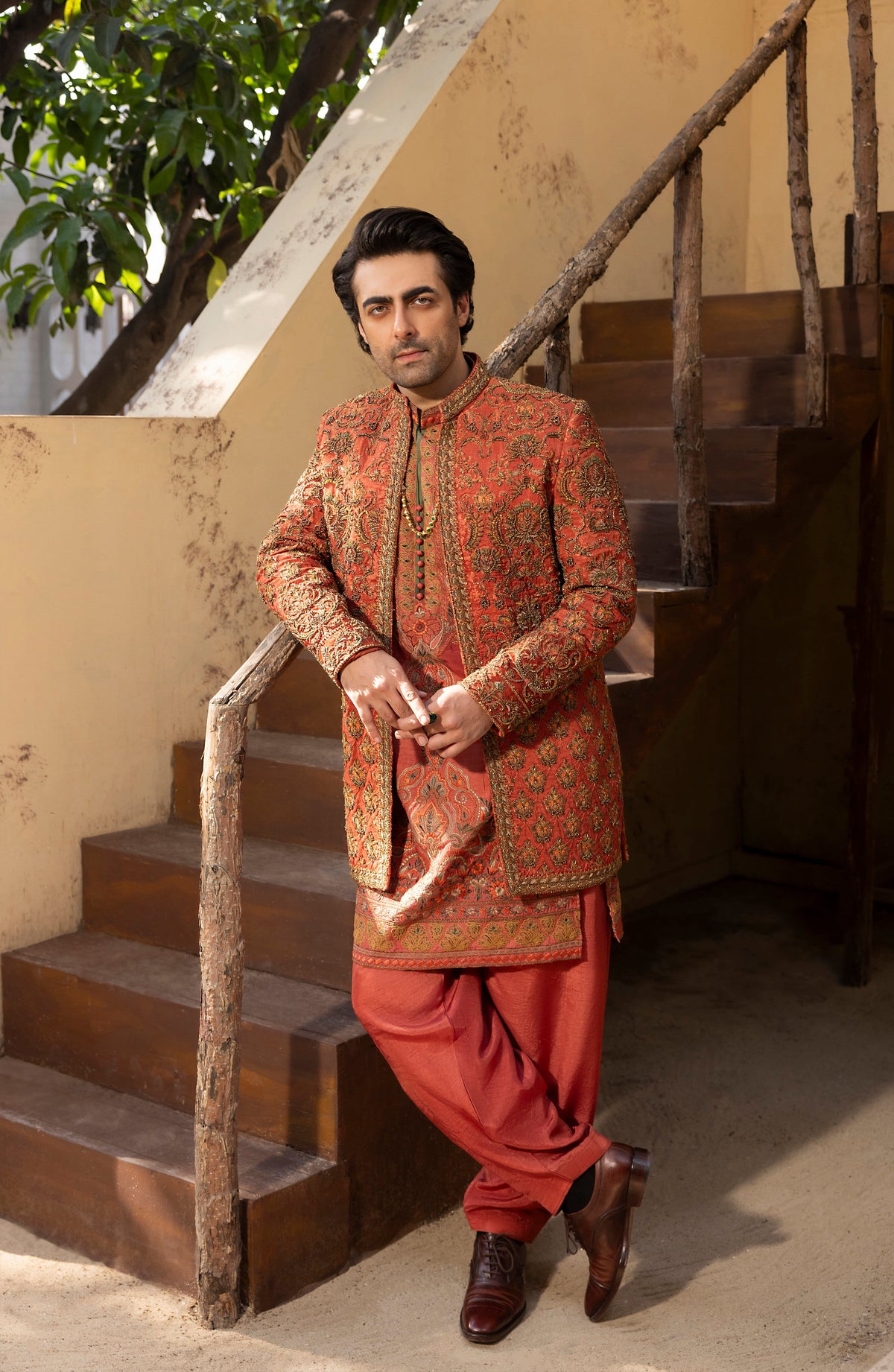 A man wearing a traditional South Asian outfit with an ornate rust-orange and gold embroidered sherwani jacket and matching red pants, standing on wooden stairs in an outdoor setting.