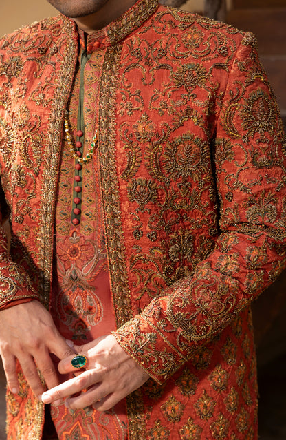 A close-up of a person wearing an ornate rust-orange sherwani jacket with intricate gold and green paisley embroidery, paired with a matching embroidered kurta and a green beaded necklace.