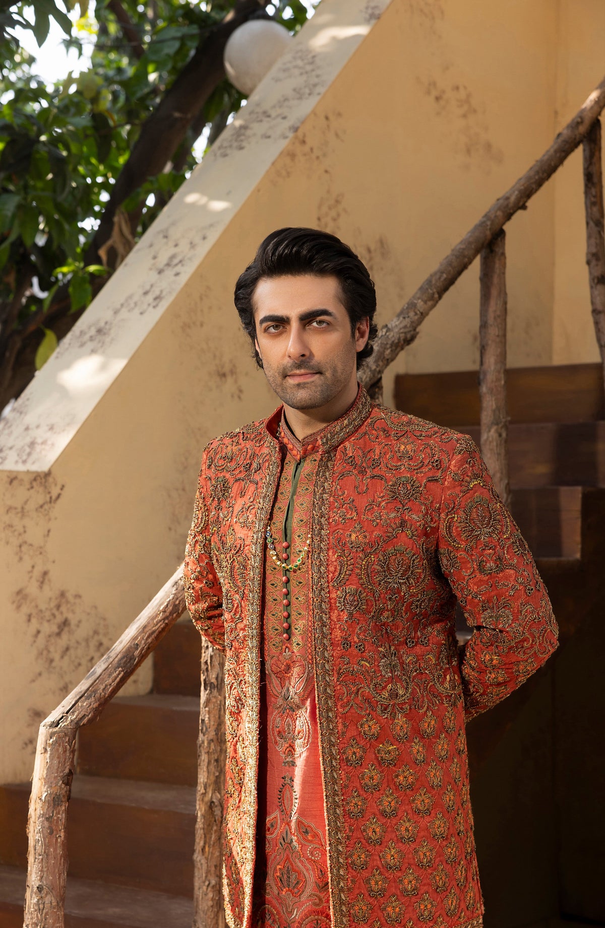 A man wearing a traditional South Asian sherwani jacket in rust orange and gold with intricate paisley patterns, standing on rustic wooden stairs against a beige wall.