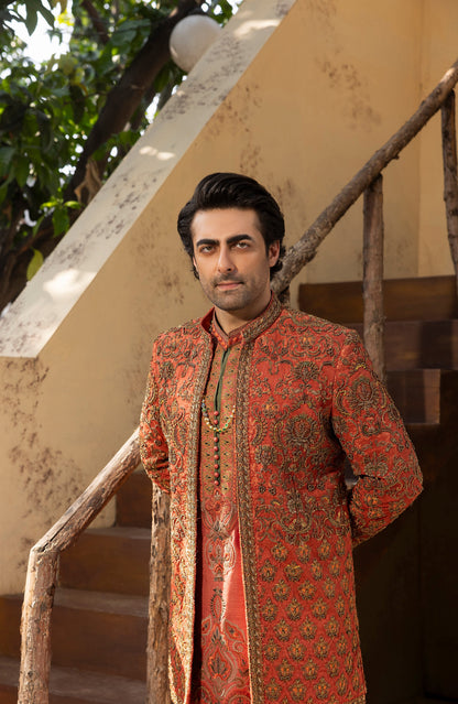A man wearing a traditional South Asian sherwani jacket in rust orange and gold with intricate paisley patterns, standing on rustic wooden stairs against a beige wall.