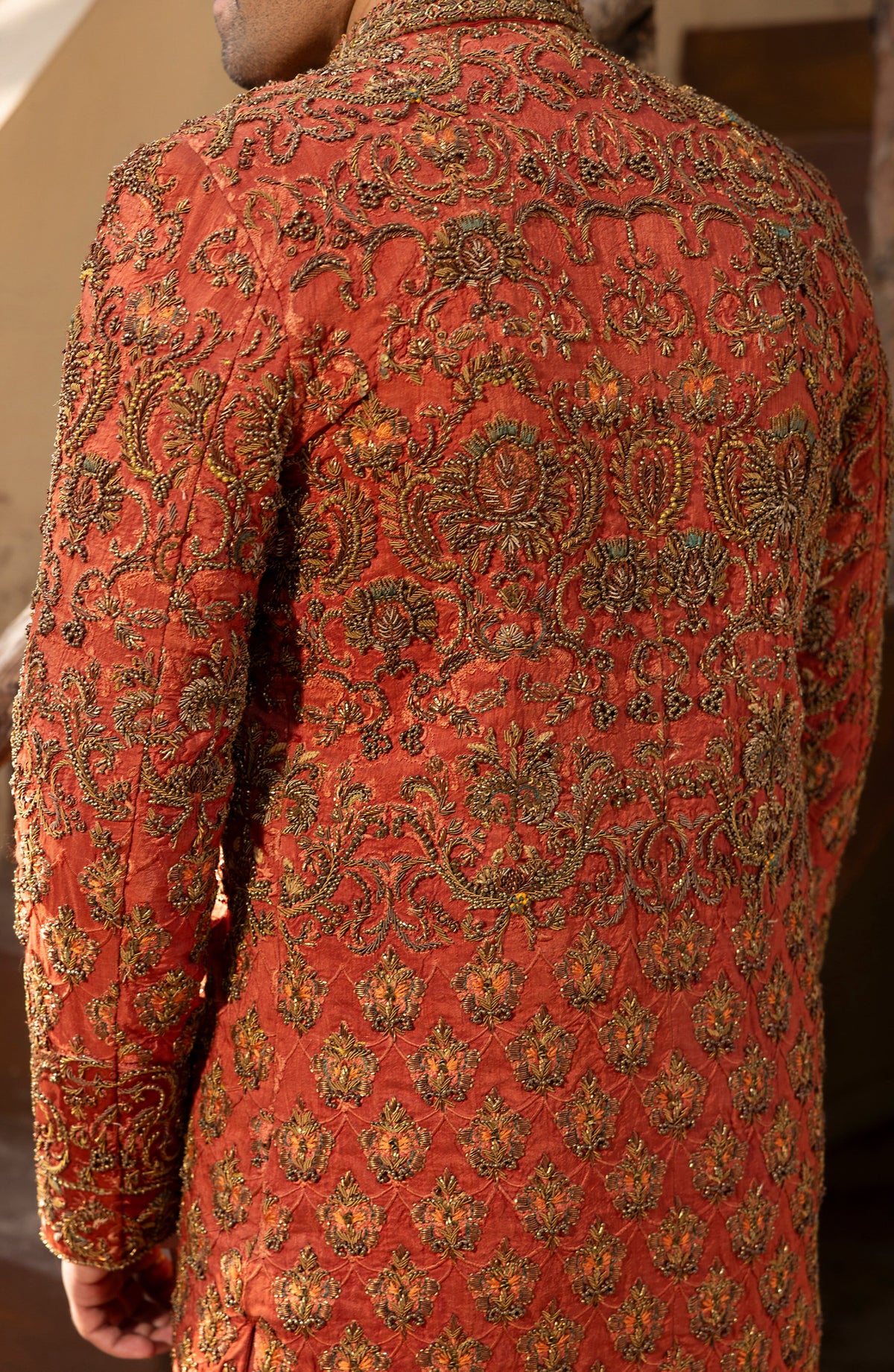 A close-up back view of a person wearing a richly embroidered orange-red sherwani jacket with intricate gold and green metallic beadwork and motifs.