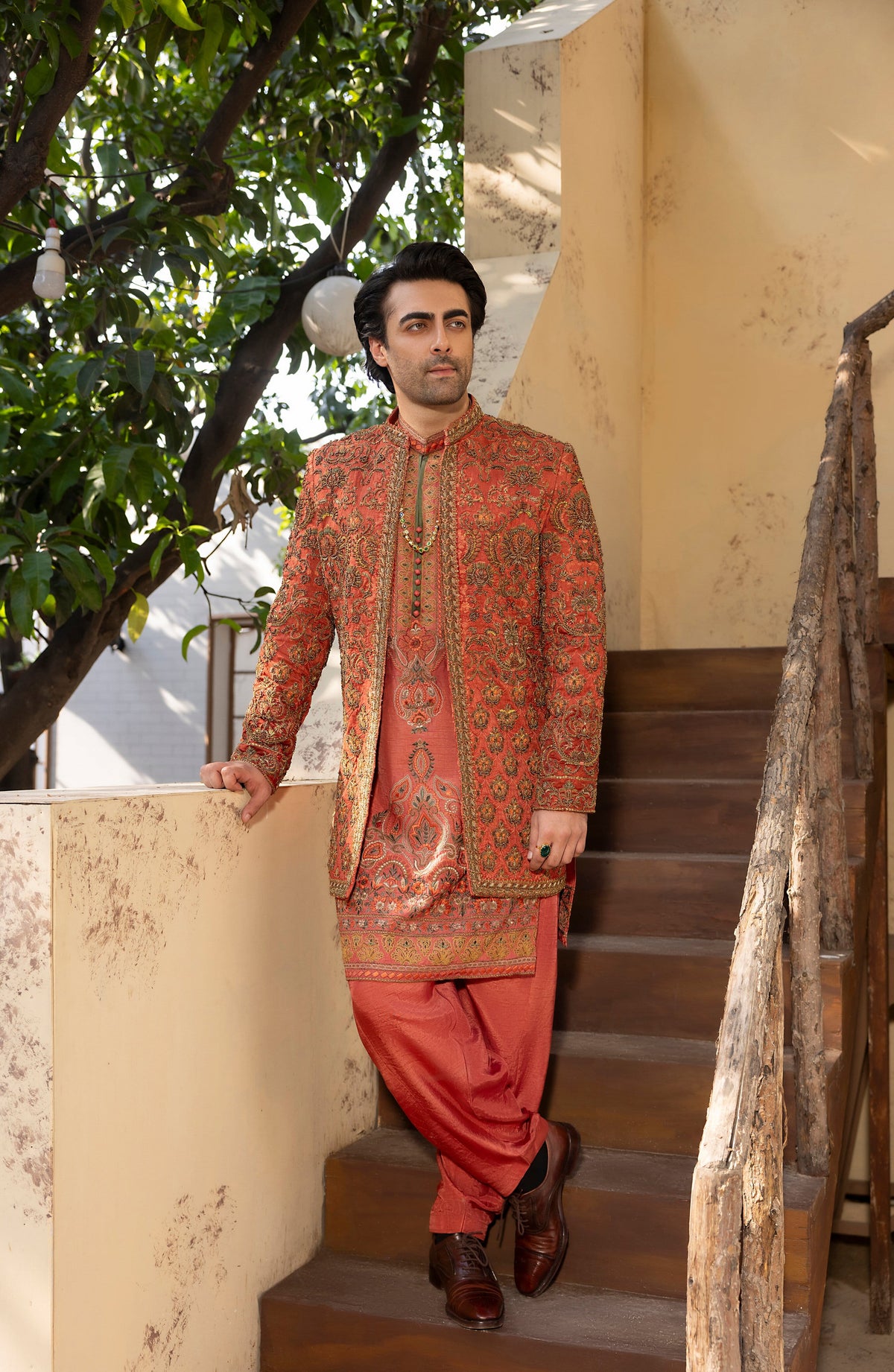 A man wearing a traditional South Asian outfit with an orange-red patterned sherwani and matching trousers, standing on outdoor wooden stairs next to a beige wall and green tree foliage.