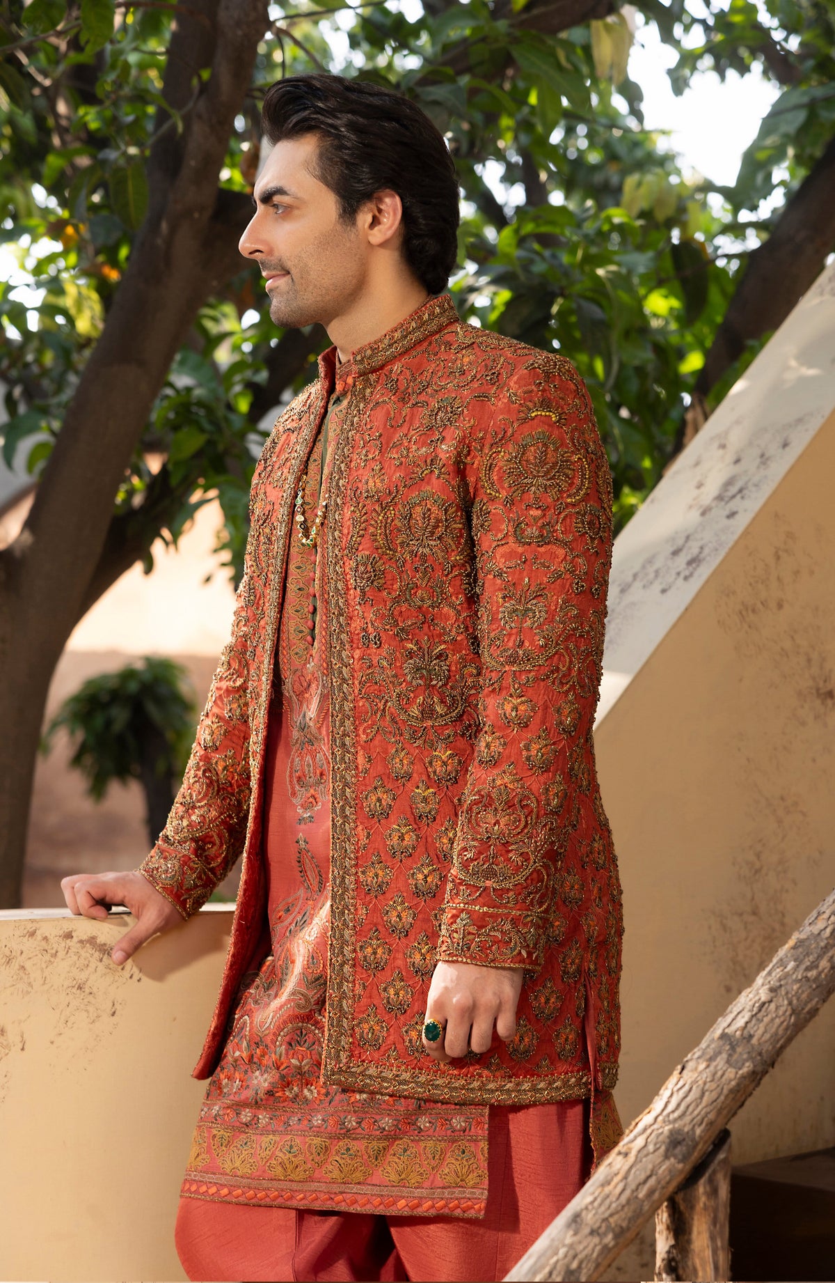 A man wearing a traditional South Asian outfit, featuring a rust-orange embroidered sherwani jacket with gold patterns over a matching rust-orange kurta and trousers, standing outdoors against a background of trees and a beige wall.