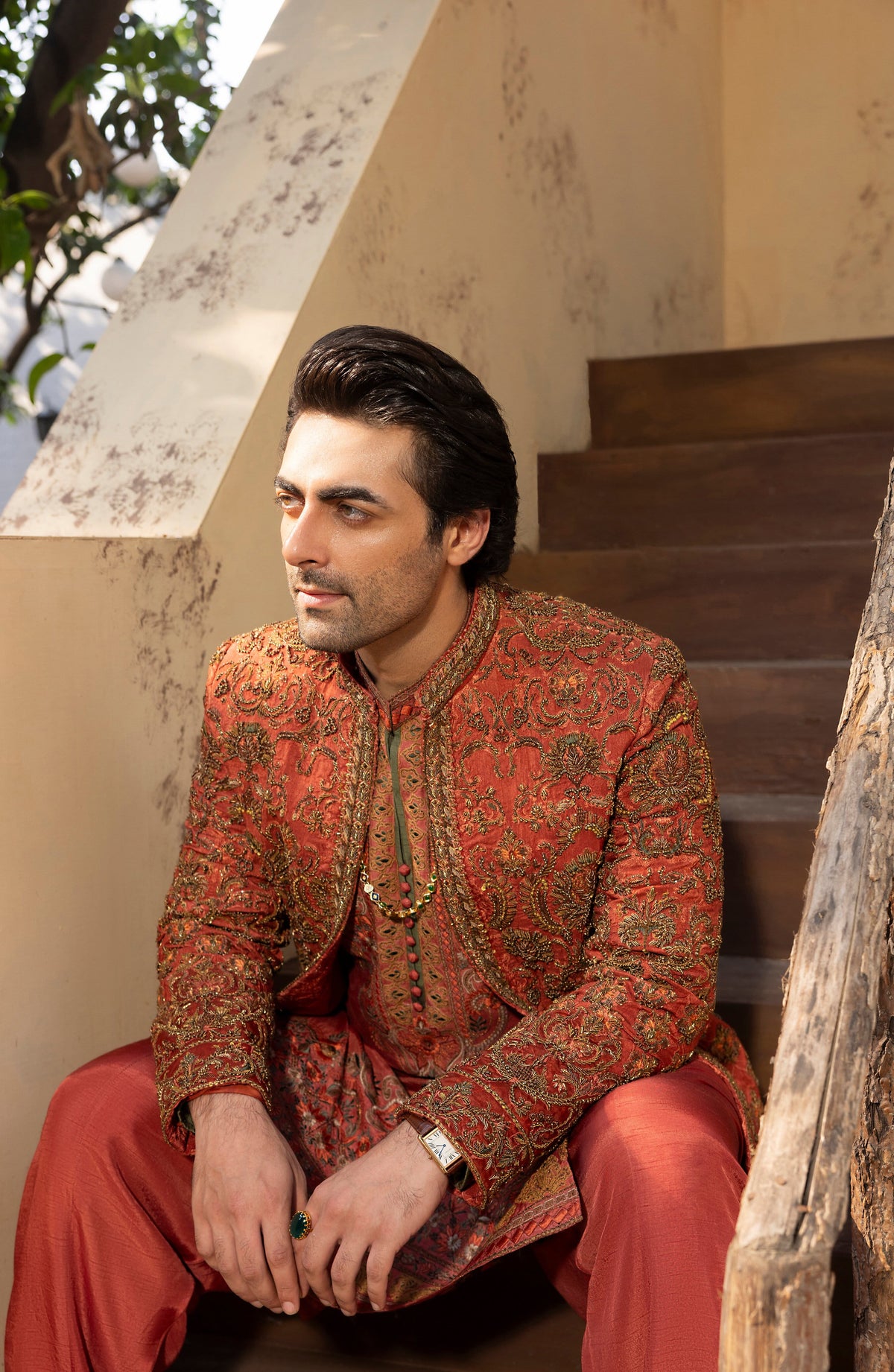 A man wearing a traditional South Asian outfit with a red and gold patterned sherwani and matching trousers, sitting on wooden stairs.