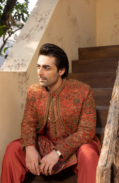 A man wearing a traditional South Asian outfit with a red and gold patterned sherwani and matching trousers, sitting on wooden stairs.
