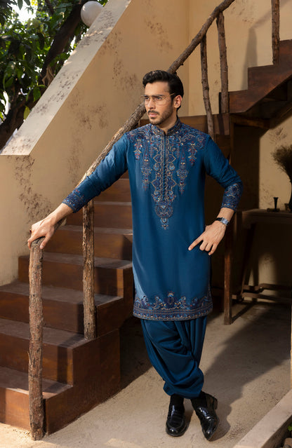 A man wearing a blue embroidered kurta and matching pants stands on a rustic outdoor staircase, holding the railing with one hand.