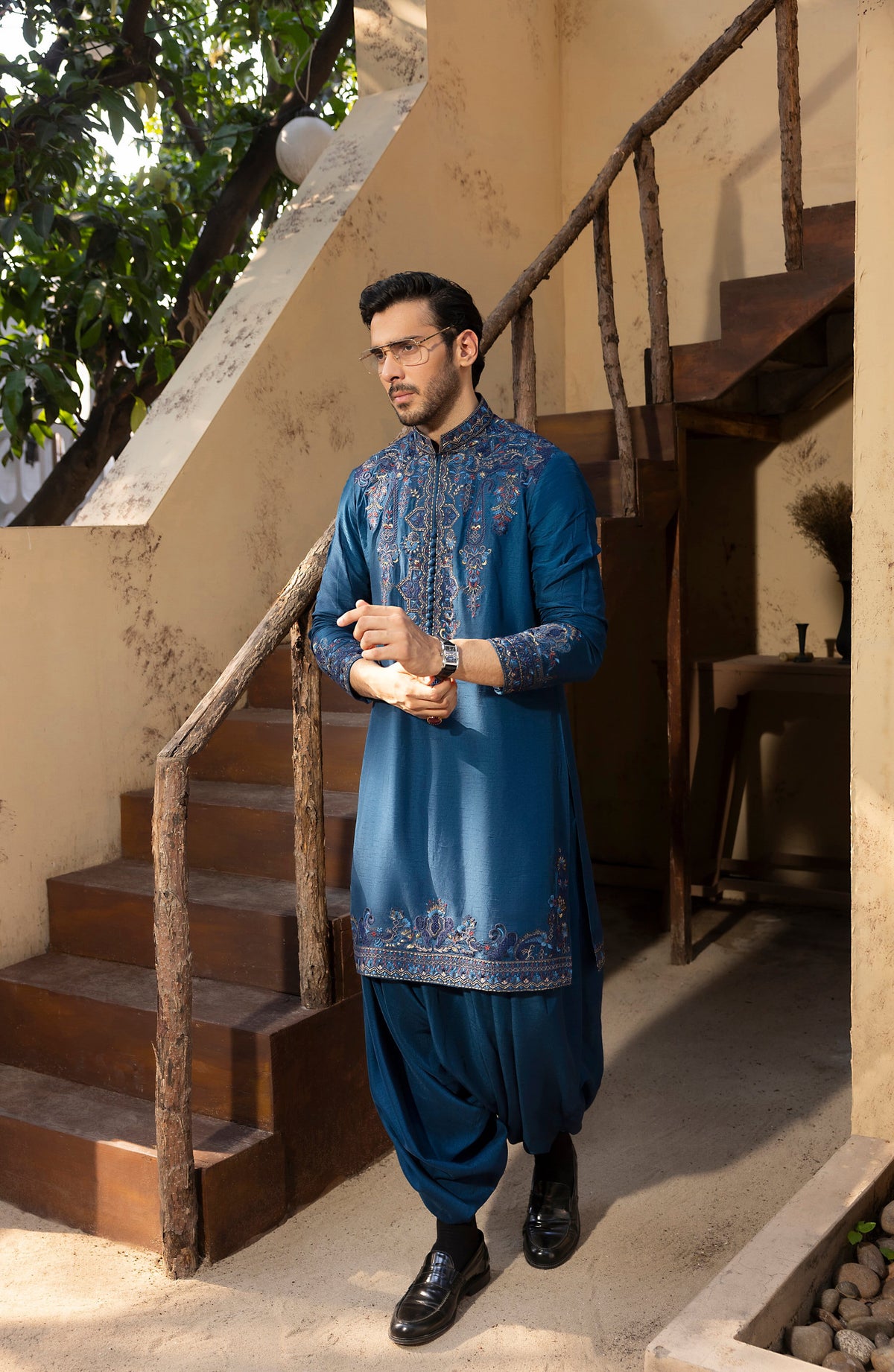 A man wearing a blue embroidered kurta and matching pants stands on a staircase landing in a warm-toned building setting.