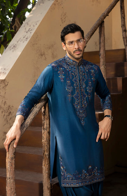 A man wearing a blue embroidered kurta and matching trousers, leaning on a rustic wooden staircase railing in an outdoor setting.