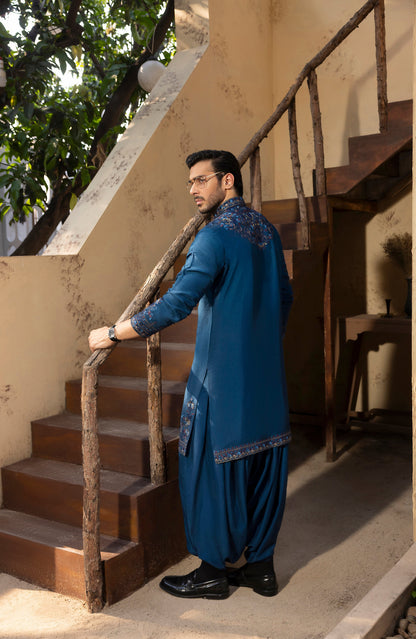 A man wearing a traditional blue kurta and pant set with embroidered details, standing on a rustic wooden staircase in a beige-walled outdoor setting.
