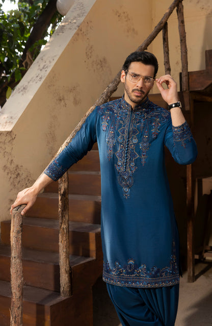 A man wearing a blue embroidered kurta and matching pants stands on rustic wooden stairs, holding the railing with one hand and adjusting his glasses with the other.
