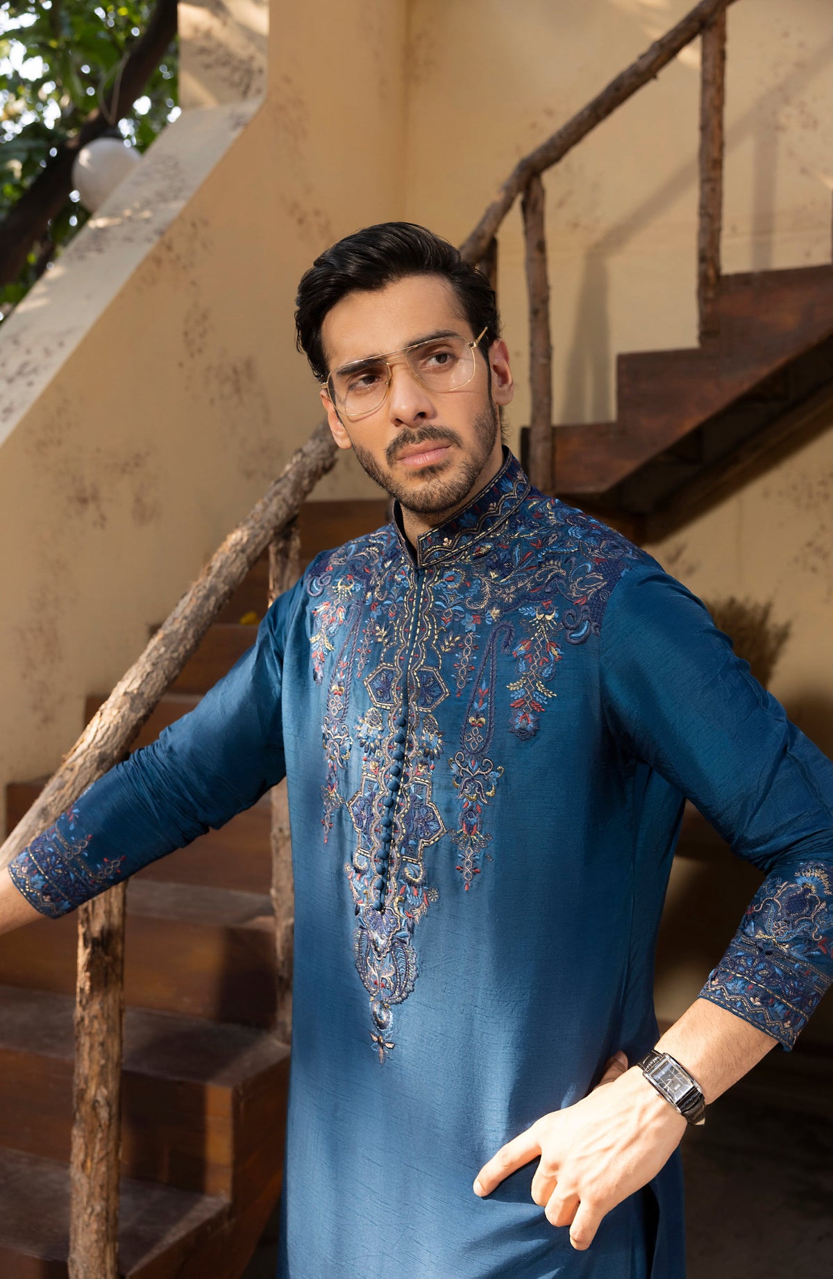 A man wearing a blue embroidered kurta stands in front of a rustic wooden staircase. The kurta features intricate silver and gold embroidery on the chest and cuffs, and he is wearing thin-framed glasses.