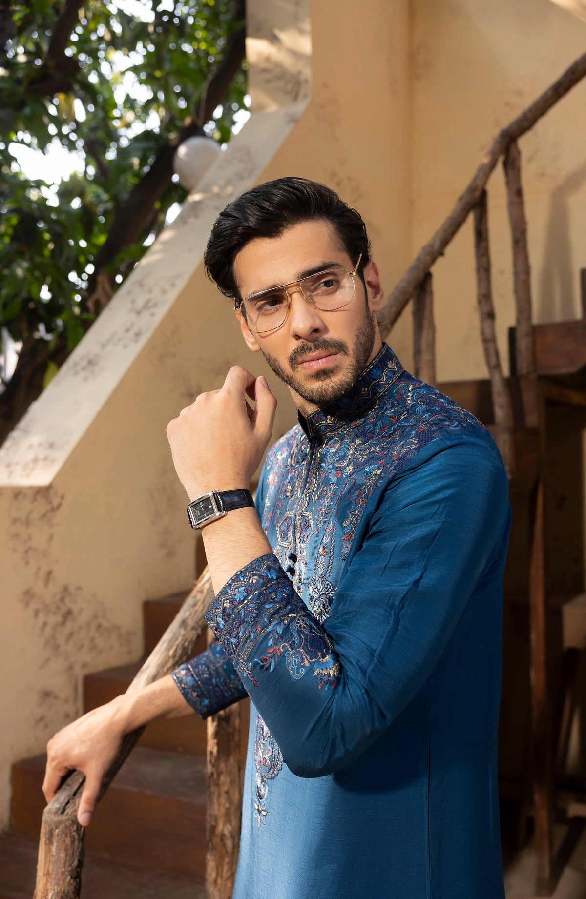 A man wearing a blue patterned kurta and glasses stands on rustic wooden stairs in a warm-toned indoor setting.