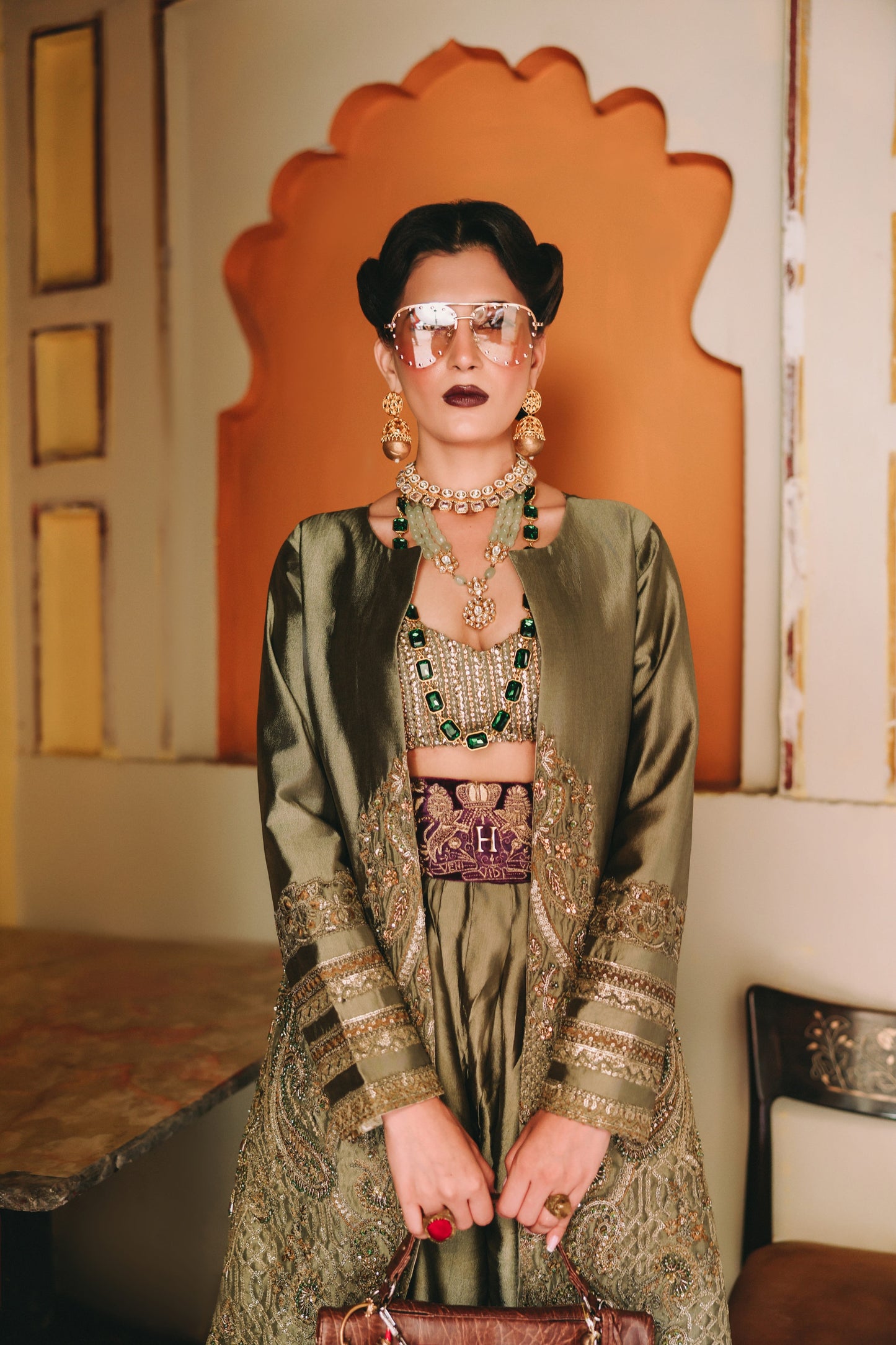 Woman in an ornate green outfit with jewelry and sunglasses in a decorated room.