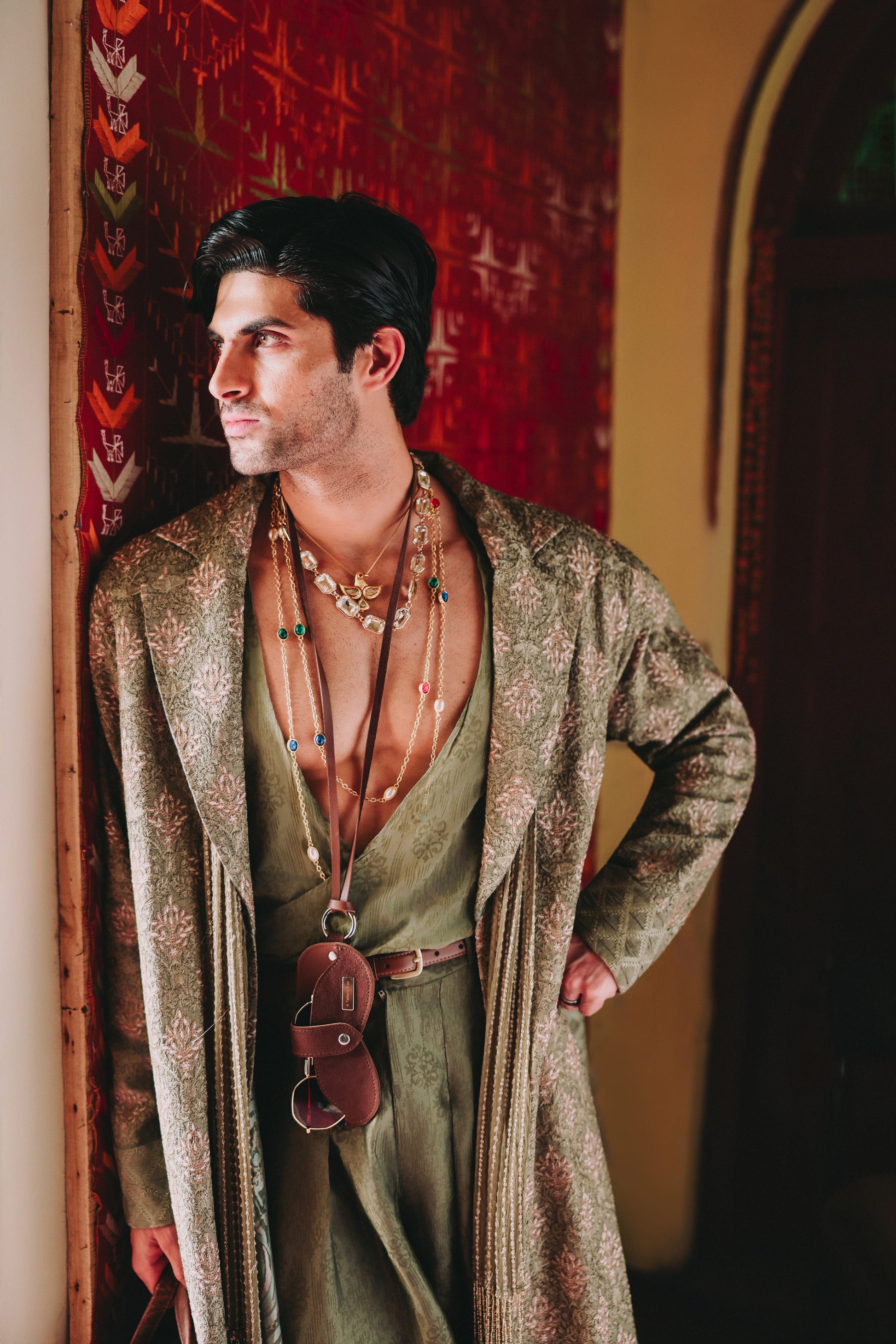 Man wearing a long, patterned robe with a red wall and archway in the background