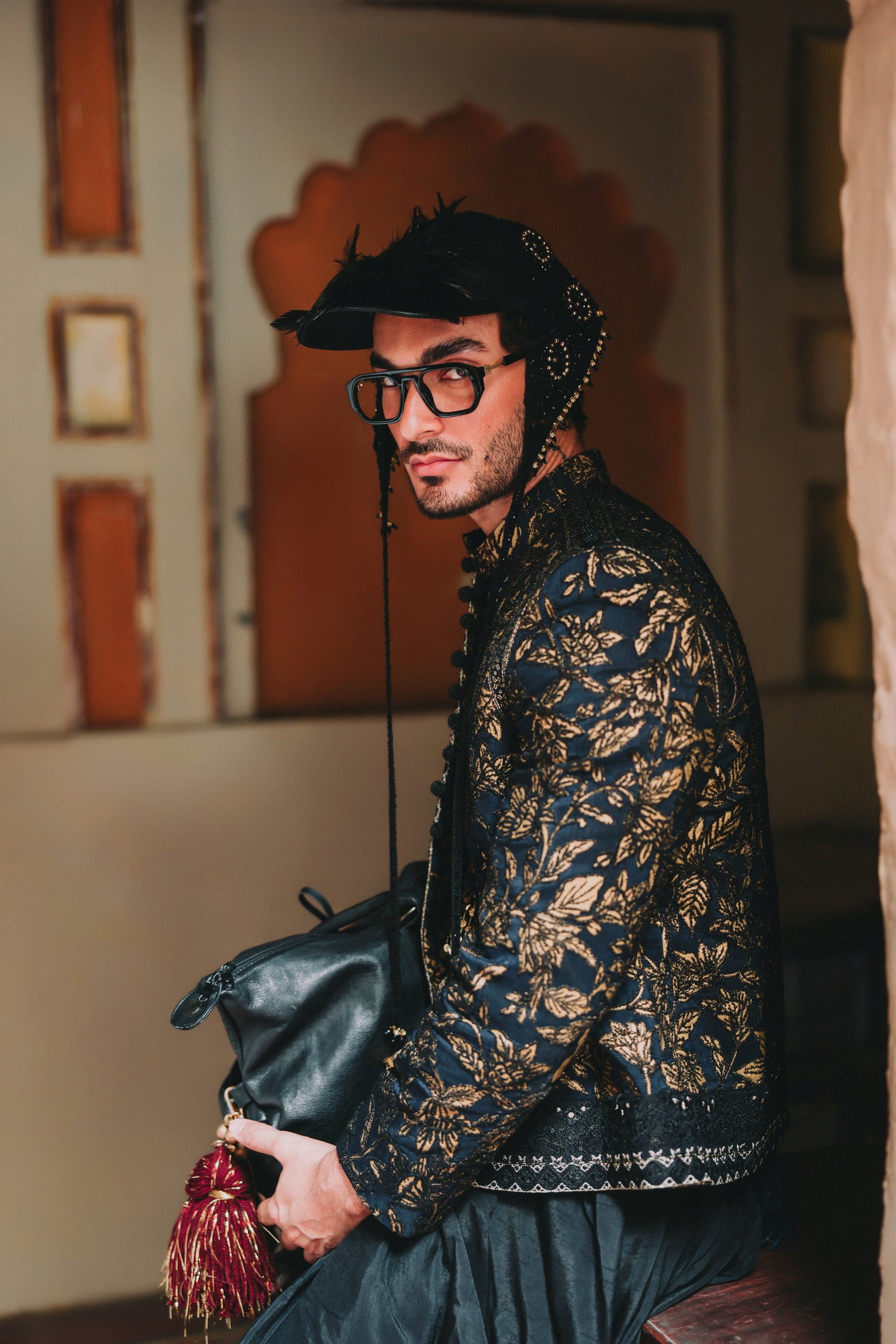 Man in traditional attire with a decorative bag, standing against an ornate wall.