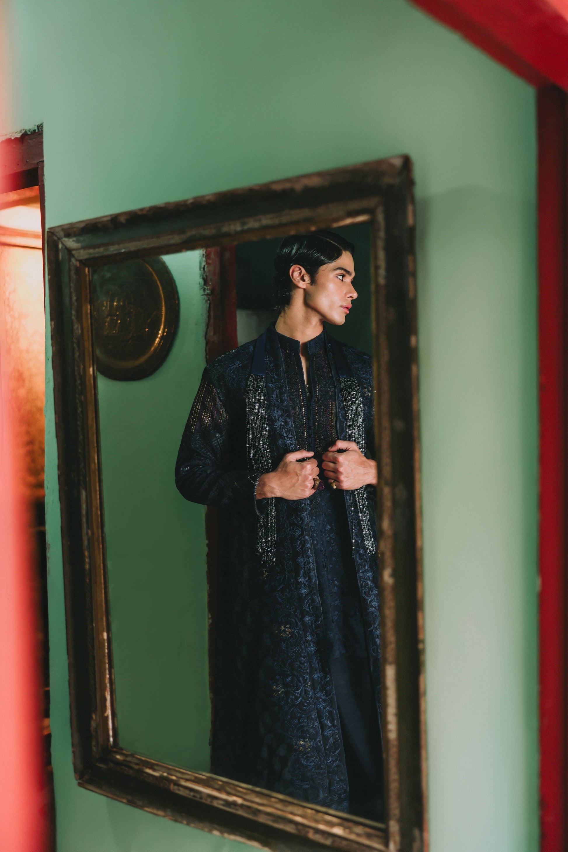 Person wearing a dark patterned outfit standing in front of a mirror with a green wall.