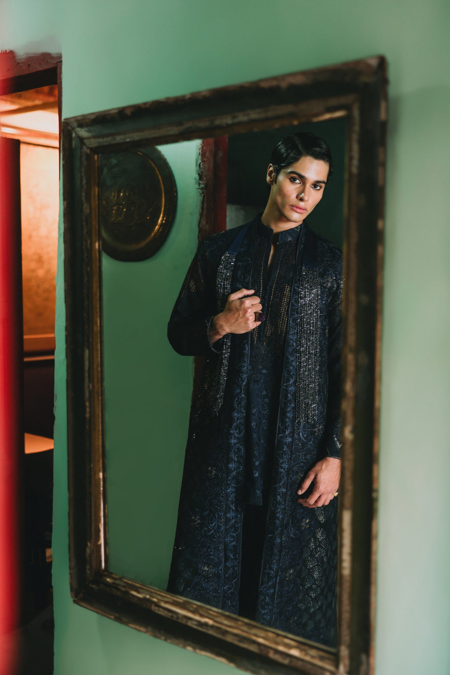 Person wearing a dark blue embroidered outfit standing in front of a vintage mirror on a green wall.