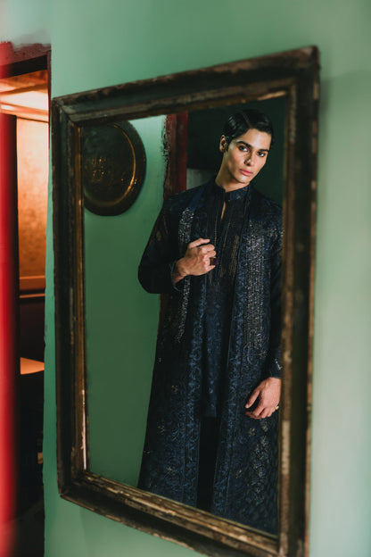 Person wearing a dark blue embroidered outfit standing in front of a vintage mirror on a green wall.