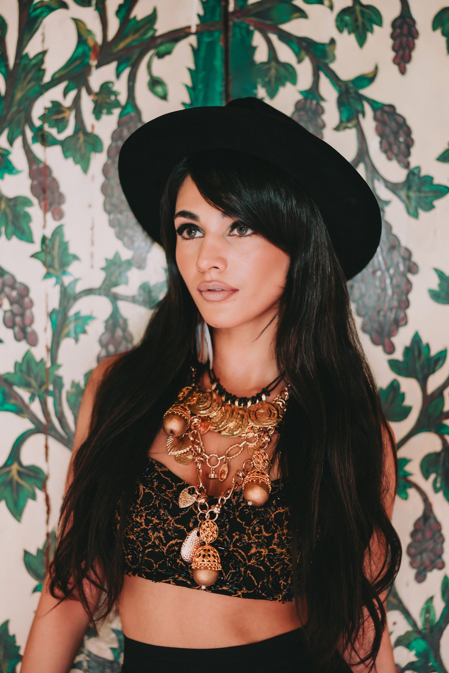 Woman wearing a black hat and gold jewelry against a decorative wall.
