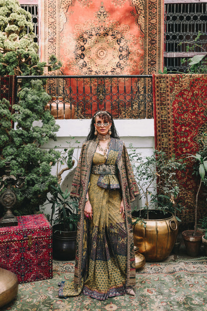 Woman in traditional attire standing in a decorative interior with patterned rugs and plants.