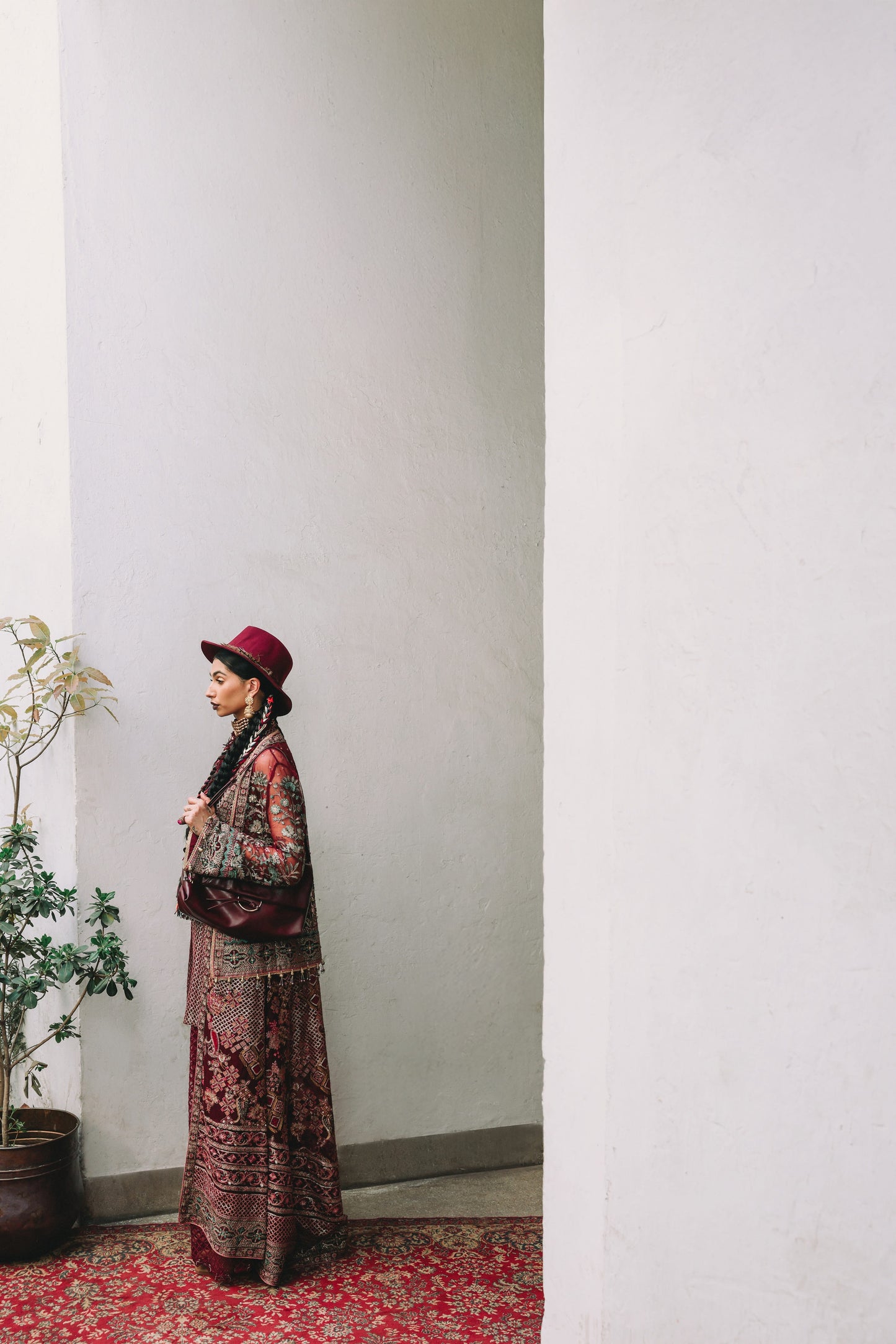 Person wearing a patterned outfit and red hat standing against a white wall with a plant to the left.