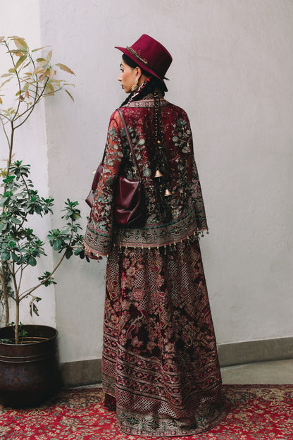 Person wearing a patterned outfit and red hat standing against a white wall with a plant to the left.