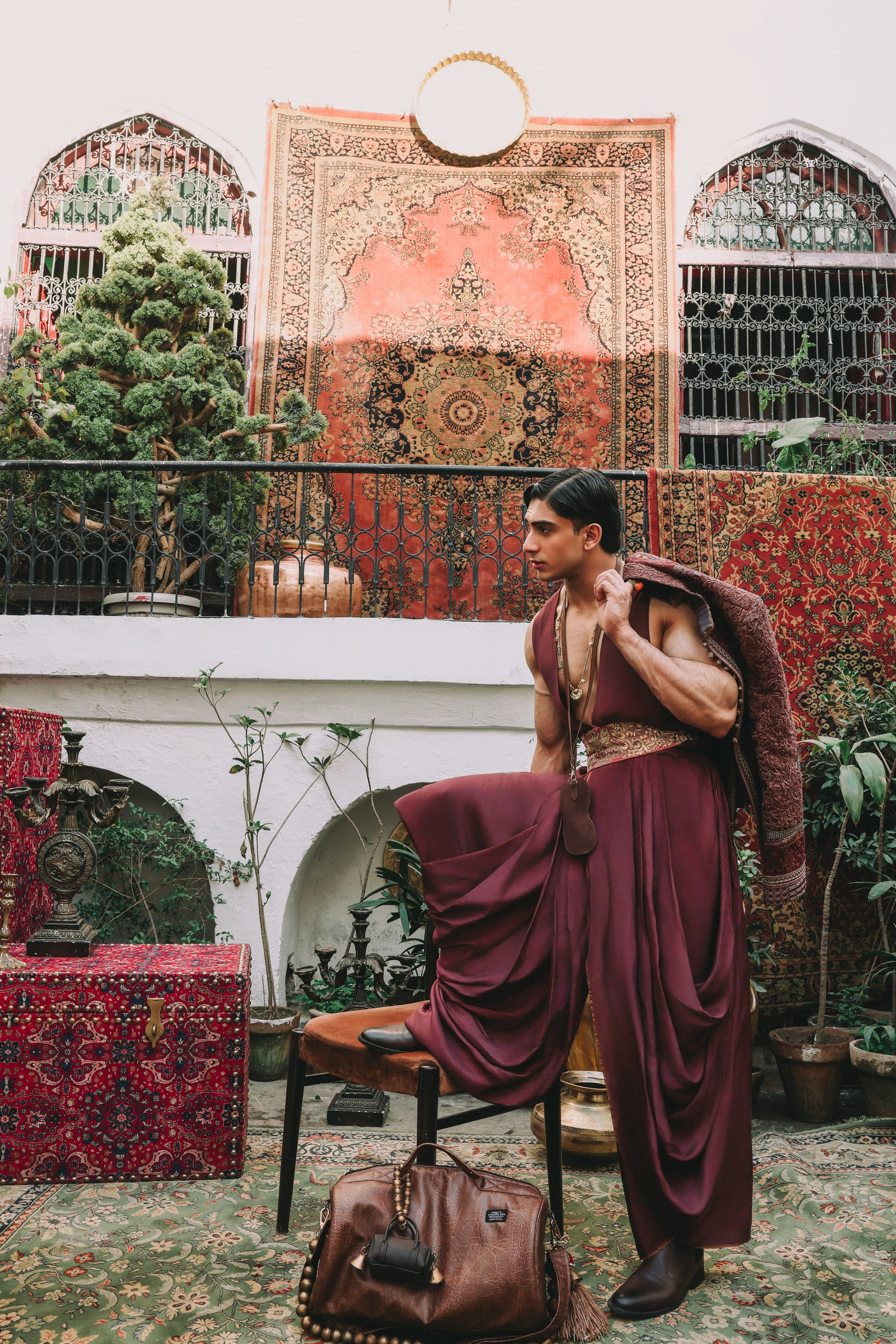 Person in a burgundy outfit sitting on a chair with a colorful rug and plants in the background