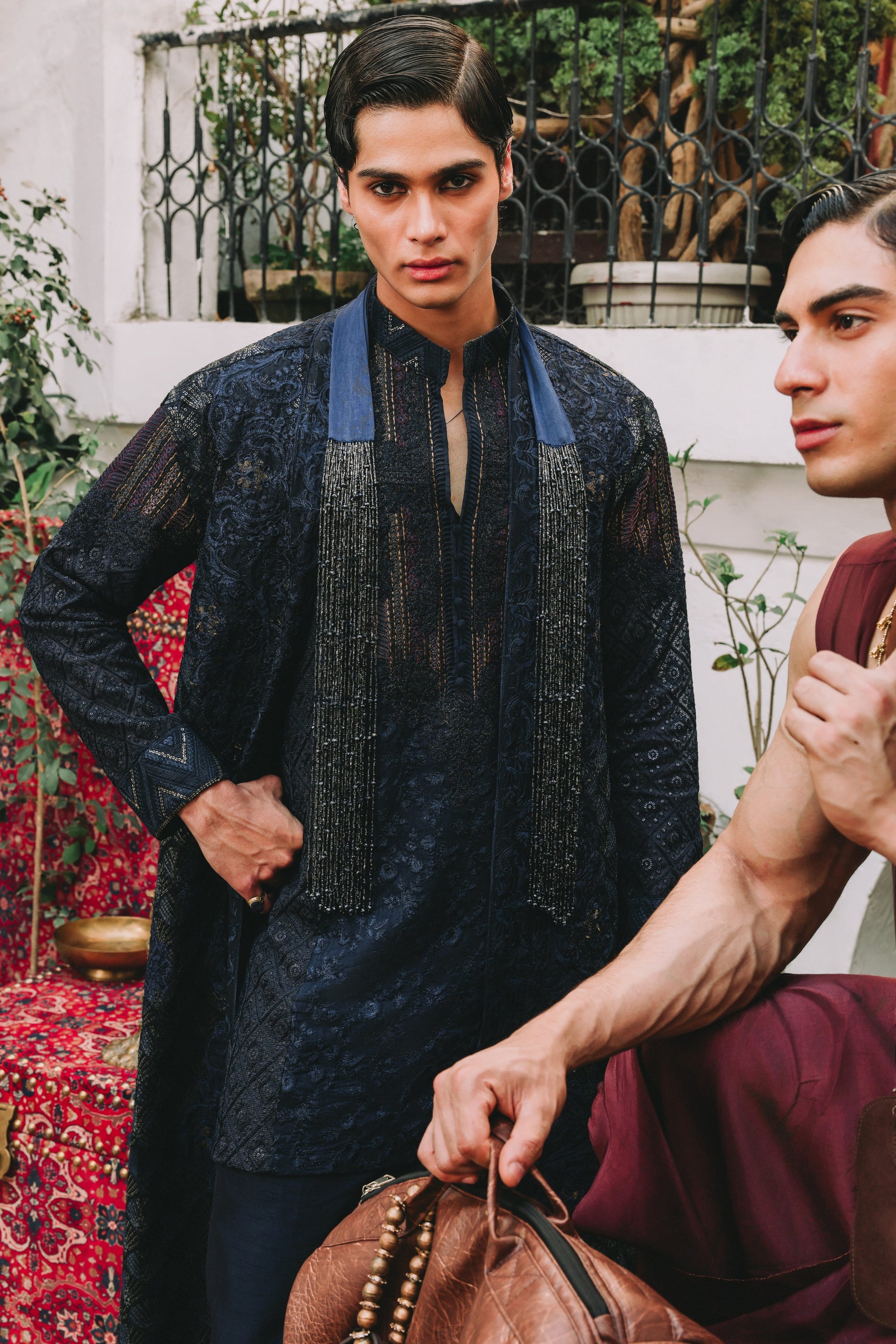 Man wearing a dark blue traditional outfit with another person holding a bag in an outdoor setting.