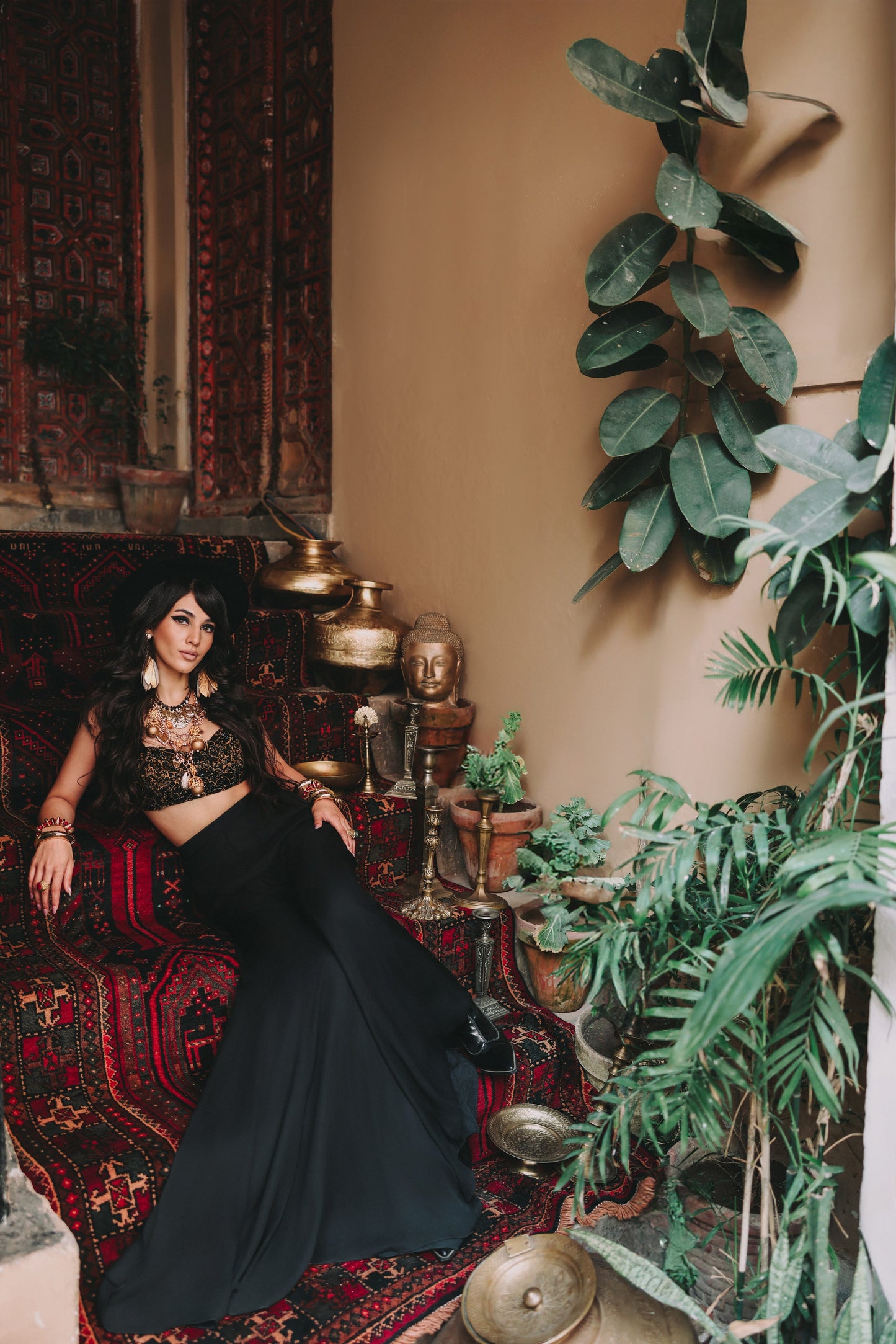 Woman in a black outfit sitting on a patterned rug with plants and decorative items around her.