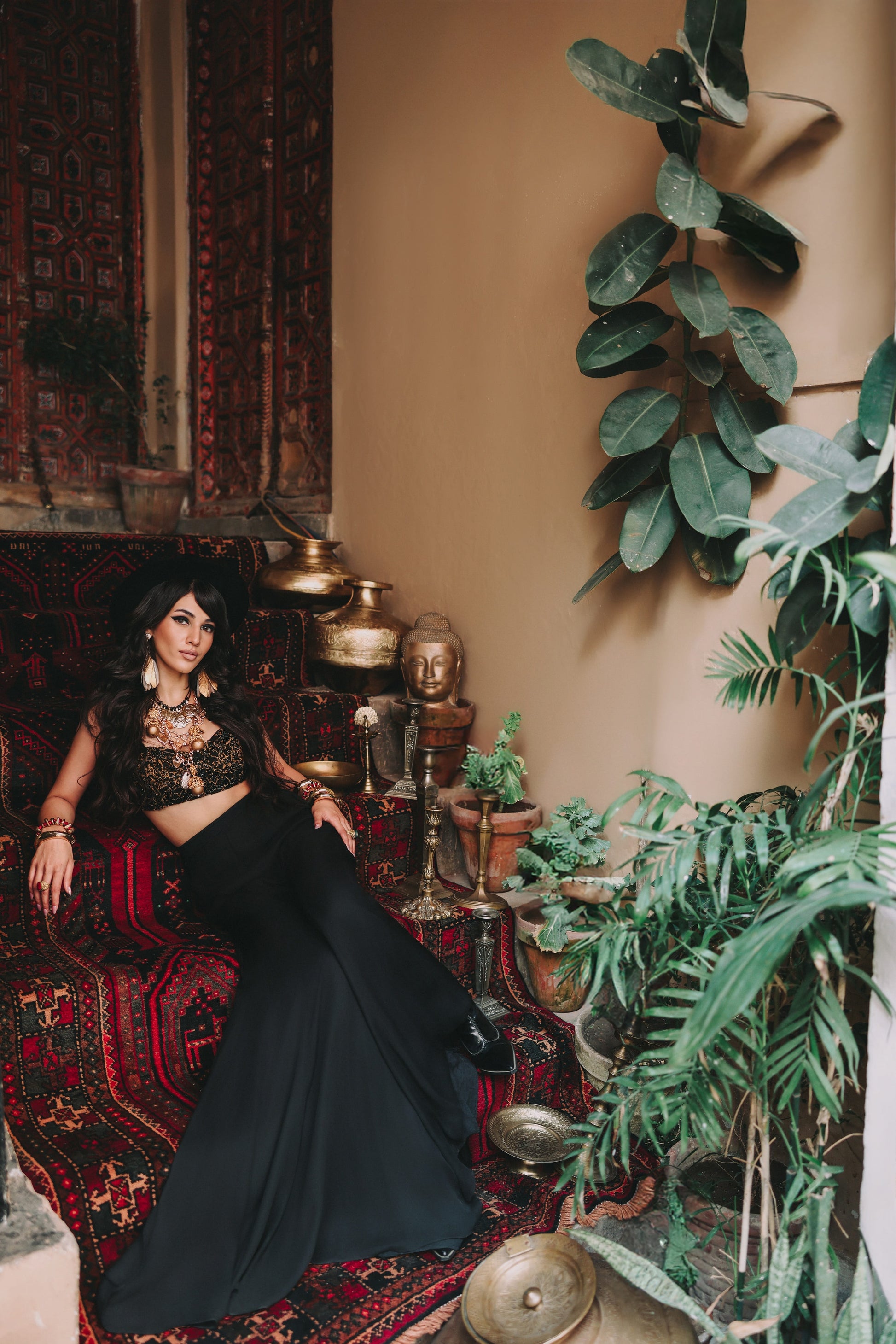 Woman in a black outfit sitting on a patterned rug with plants and decorative items around her.