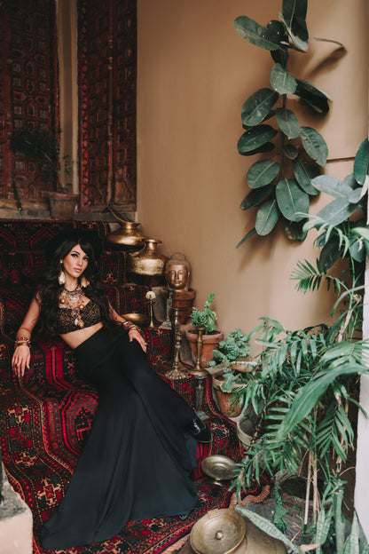 Woman in a black outfit sitting on a patterned rug with plants and decorative items around her.