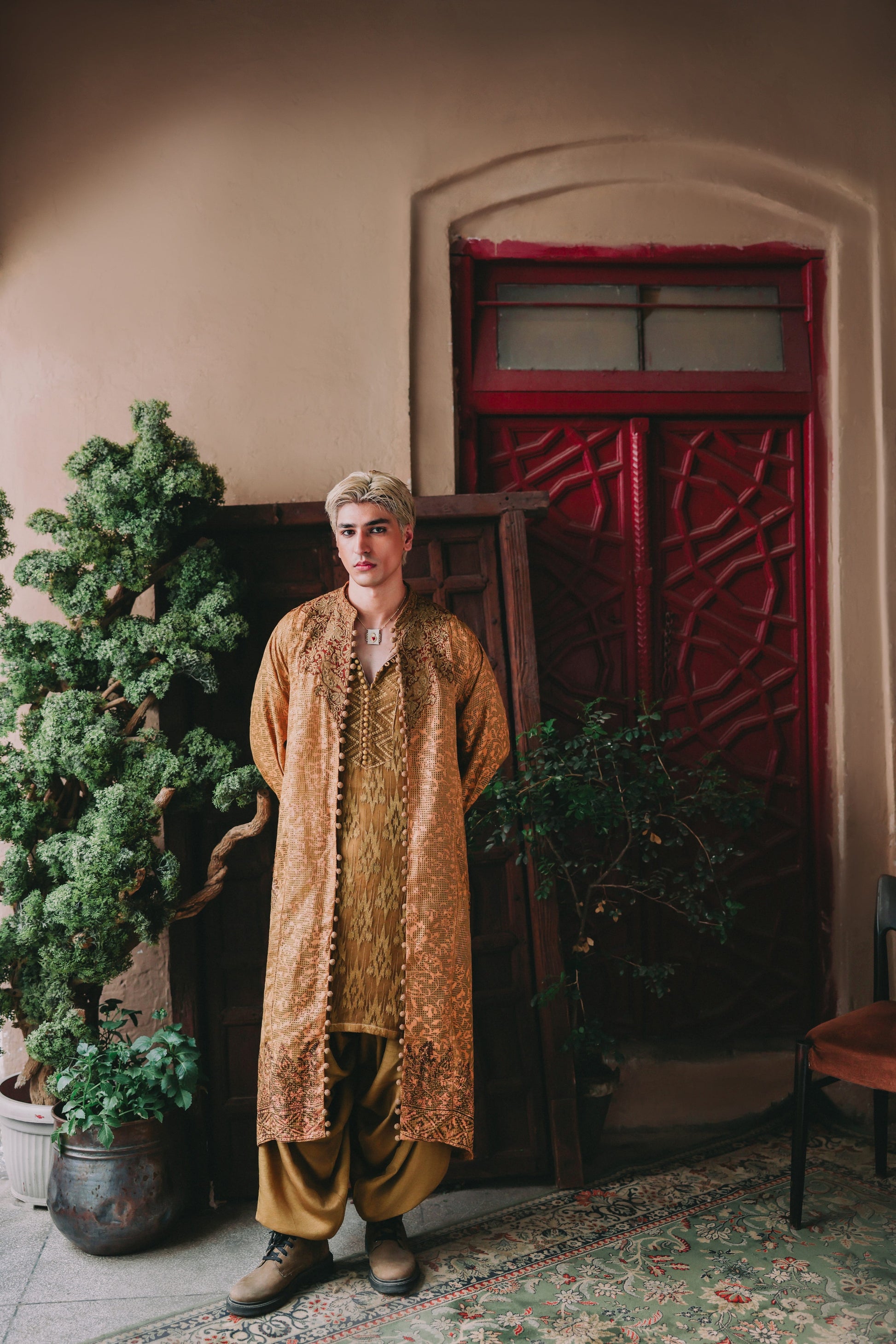 Person wearing a patterned outfit standing in a room with a red door and plants.