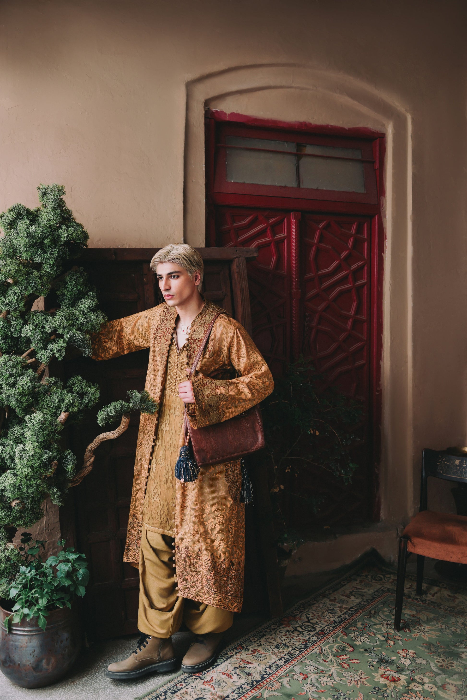 Man in a long, patterned coat standing in a room with a red door and plants.