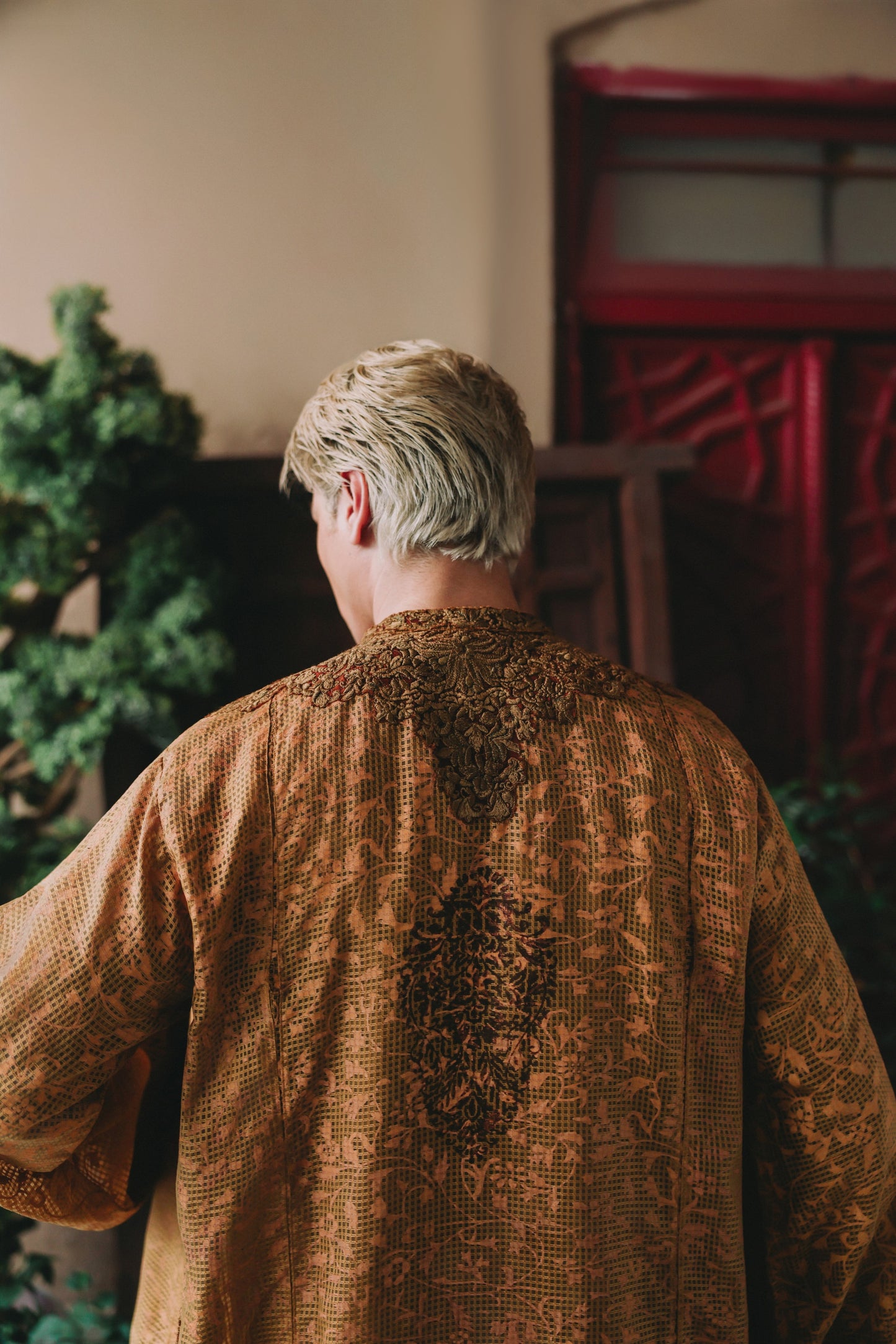 Person wearing a textured brown garment standing in a room with a red door and greenery.